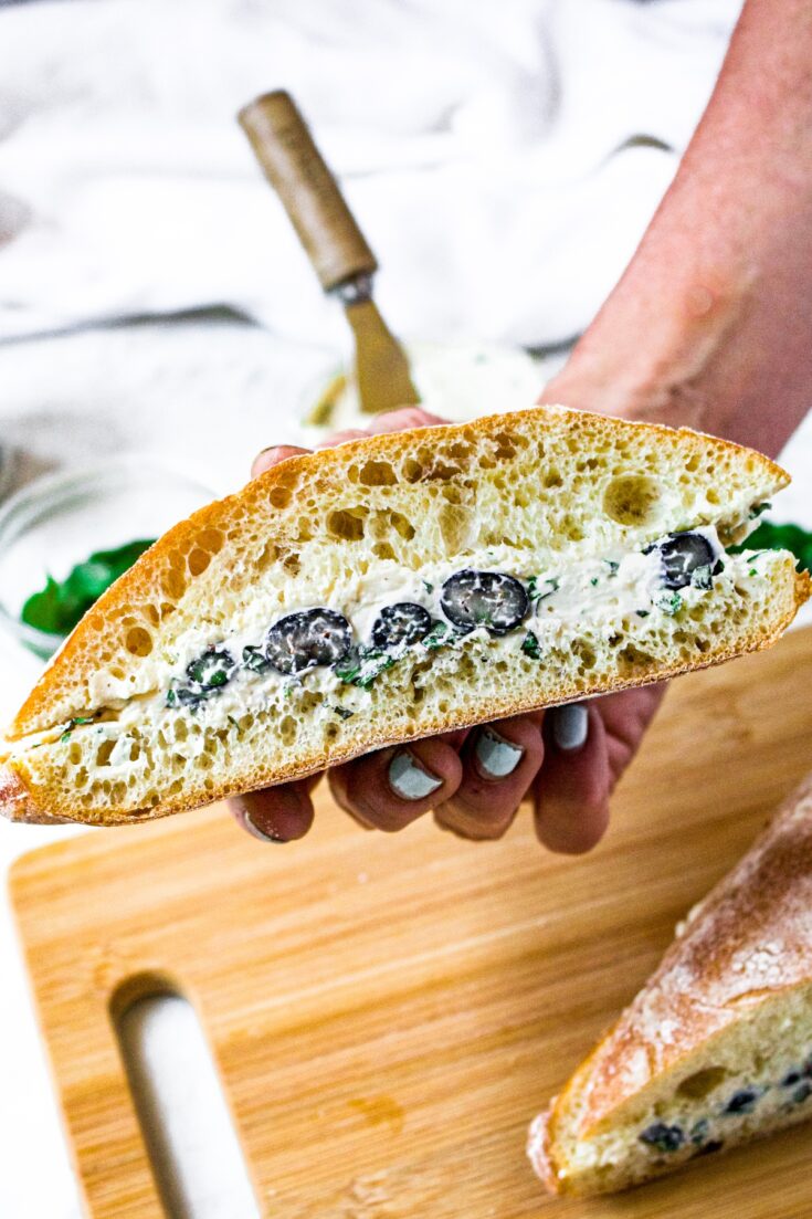A hand holding half a ciabatta sandwich with creamy vegan goat cheese, fresh blueberries, and minced basil.