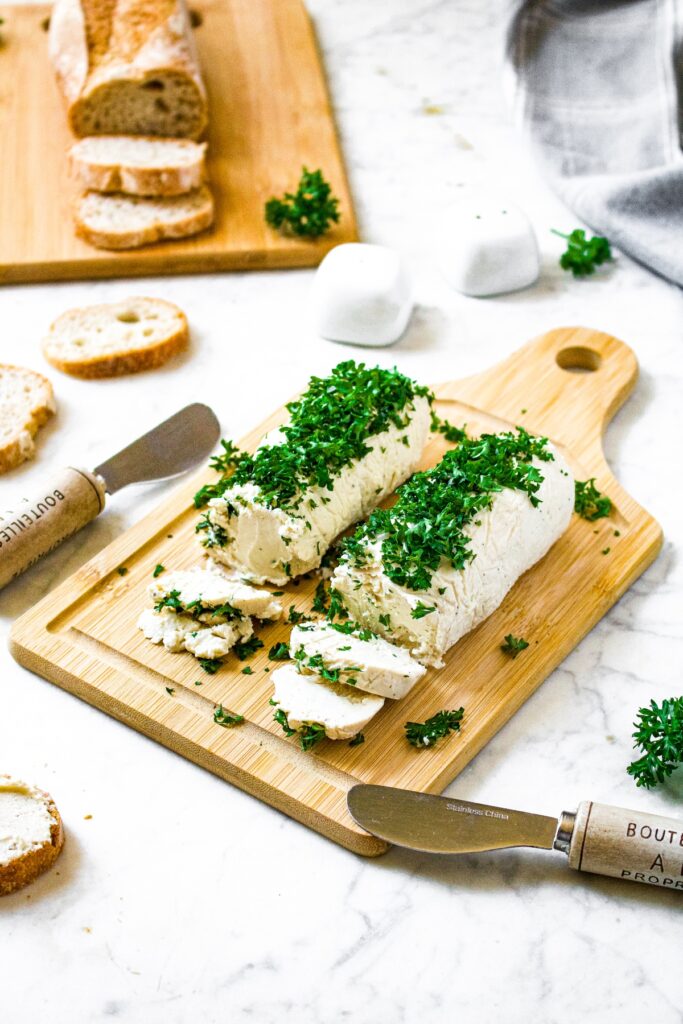 Head-on shot of two tubes of vegan goat cheese on a wooden cutting board topped with fresh minced parsley. There are slices of baguette surrounding the board along with cheese knives, and salt and pepper shakers