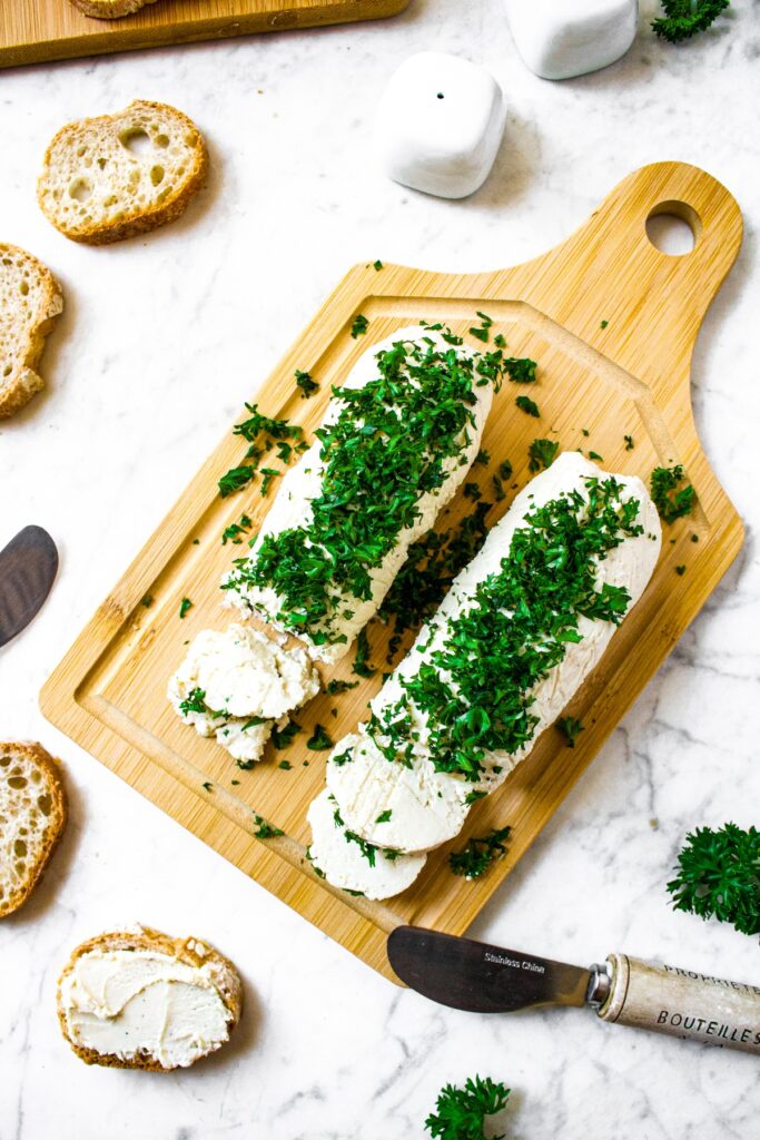 Overhead shot of 2 logs of dairy free goat cheese topped with fresh minced parsley that is very green. The tofu cheese sits on top of a wooden cutting board and is surrounded by cheese knives and slices of bread. One slice of bread has tofu cheese spread on it.