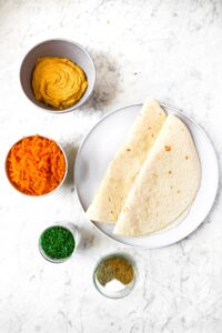 Overhead shot of the ingredients you need to make our vegan pinwheel recipe: flour tortillas, hummus, grated carrots, chopped chives, salt, pepper, and ground coriander.