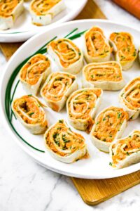 Overhead shot of a large round plate with hummus pinwheels on it. There is a fresh chive on the edge of the plate and another plate behind it.