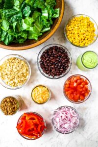 Overhead shot of all the ingredients you need to make a vegetarian southwest salad: greens, corn, black beans, diced tomato, red onion, red pepper, avocado dressing, vegan cheese shreds, walnut chorizo, and crushed tortilla chips
