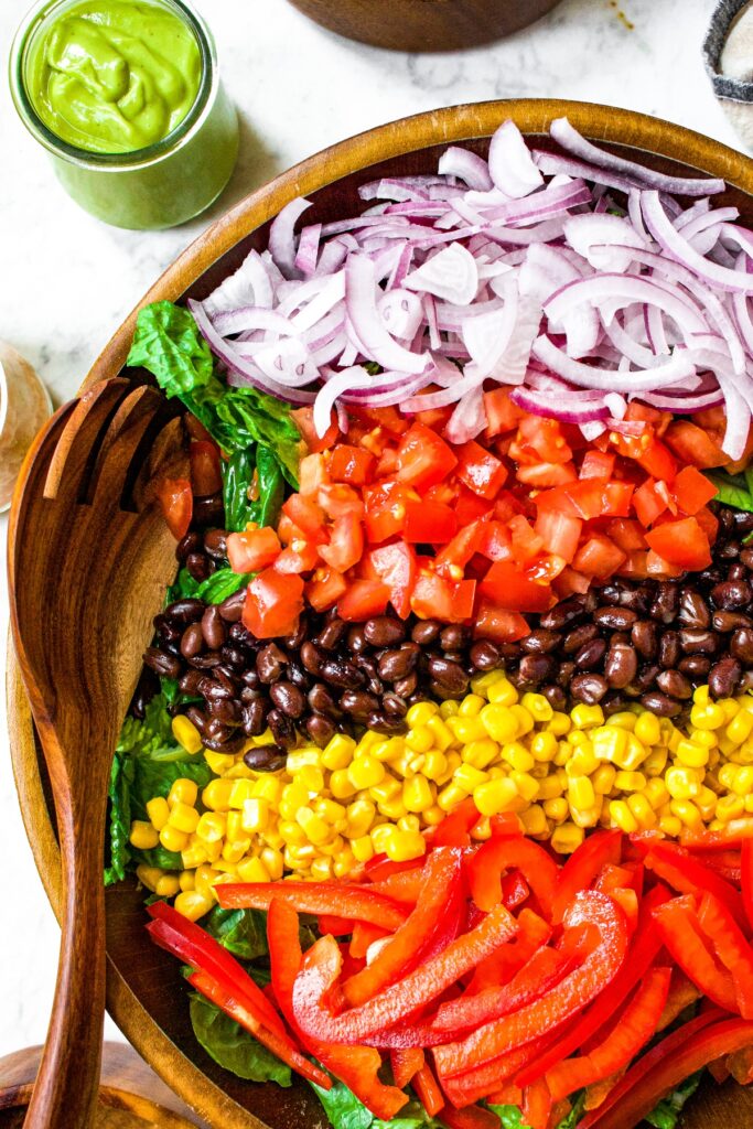 Overhead shot of a large bamboo salad bowl with greens, corn, red peppers, tomato, red onion, and black beans segmented in the bowl.