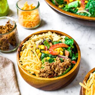 Overhead shot of a small bamboo salad bowl filled with a green southwest salad topped with dairy free cheddar, vegan chorizo, and tortilla chips. There is a larger salad bowl in the background along with small clear jars of shredded cheddar, walnut chorizo, and avocado dressing.