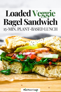 Close up head on shot of a garden vegetable bagel sandwich sliced in half to display the layers of veggie fillings: spinach, tomato, cucumber, red onion, pickles, and roasted red peppers. Text reads: loaded veggie bagel sandwich, 15-min. plant-based lunch