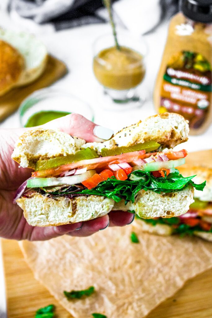 Close up overhead shot of a hand holding a vegan bagel sandwich that has been cut in half. A bottle of Organicville stone ground mustard is in the background with a small clear bowl filled with mustard sits next to it.