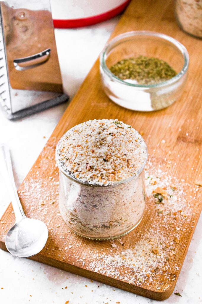 An overhead shot of a clear glass jar of Italian herb bread crumbs with a spoon next to it, cheese grater behind it, and a small clear bowl of spices and dried herbs in the background