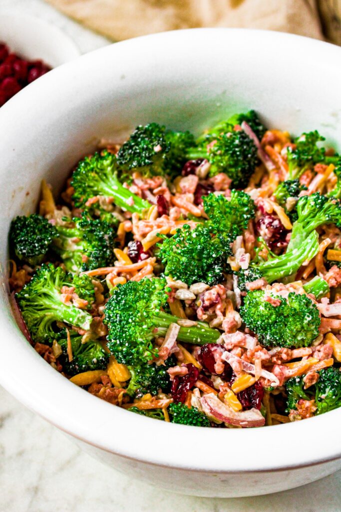 Overhead shot of a big white mixing bowl filled with vegan broccoli salad loaded with carrots, vegan cheese, crainsins, and imitation bacon bits