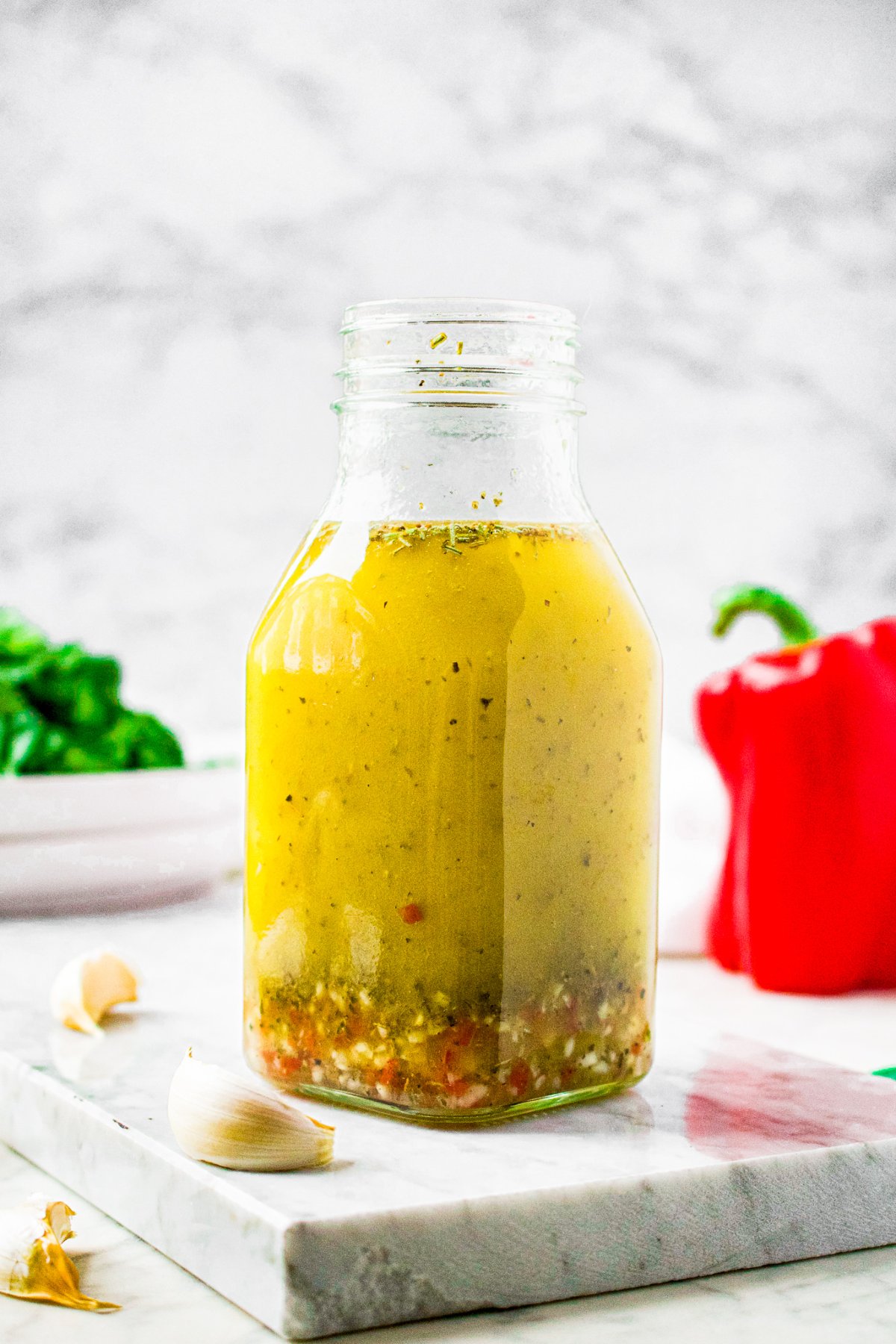Head-on shot of a bottle filled with homemade Italian salad dressing. There are garlic cloves around the bottle and a standing red bell pepper in the background.