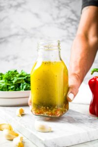 A hand reaches in and lifts up a bottle of Italian salad dressing. There is a plate of lettuce and red bell pepper in the background and garlic cloves all around the scene.