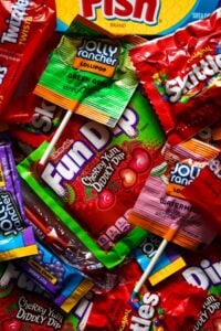 Overhead view of a pile of vegan candy like Fun Dip, Skittles, and Jolly Rancher Lollipops with a hand is reaching in to grab one of the lollipops.