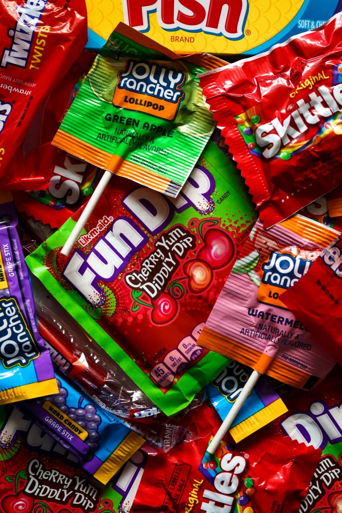Overhead view of a pile of vegan candy like Fun Dip, Skittles, and Jolly Rancher Lollipops with a hand is reaching in to grab one of the lollipops.