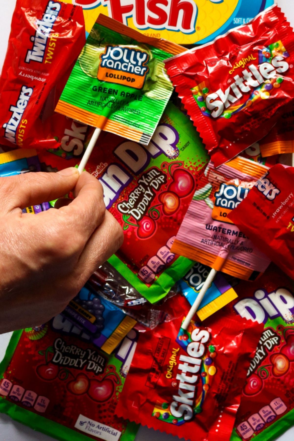 Overhead view of a pile of vegan candy like Fun Dip, Skittles, and Jolly Rancher Lollipops with a hand is reaching in to grab one of the lollipops.