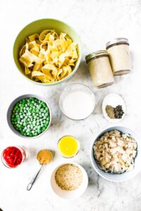 Overhead shot of all the ingredients you need to make vegan tuna noodle casserole: noodles, peas, pimentos, miso, non-dairy milk, vegan cream of mushroom, jackfruit, spices, vegan butter, and breadcrumbs