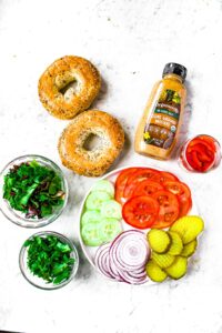 Overhead shot of the ingredients needed to make simple cold vegetarian sandwiches: bagels, mustard, greens, pickles, red onion, cucumbers, roasted red peppers, tomatoes