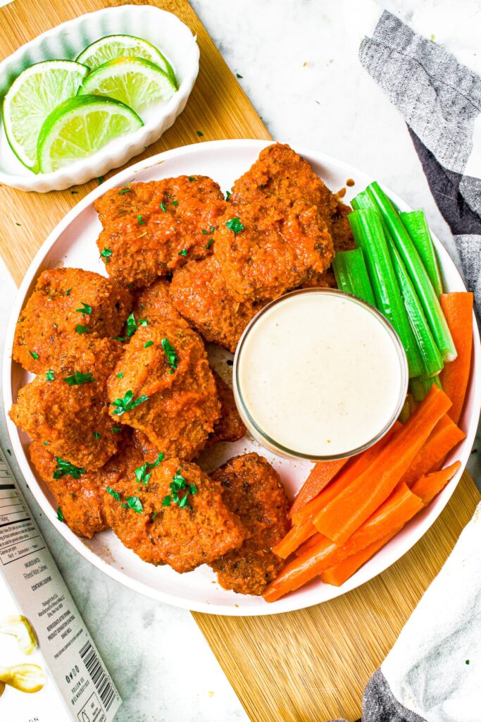 Overhead shot of a round white plate with vegan buffalo wings piled on one side, a glass cup of blue cheese in the center, and carrot sticks and celery sticks to the right side. There is an oval-shaped bowl filled with lime wedges in the upper left corner
