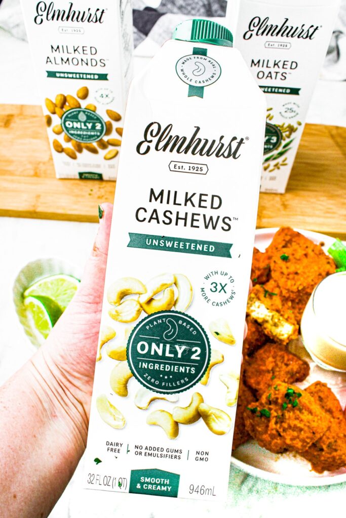 A hand holding a carton of Elmhurst unsweetened milked cashews over a plate of air fryer vegan buffalo wings