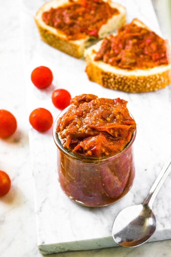 Overhead shot of a jar of chunky cherry tomato jam with cherry tomatoes behind the jar along with a slice of bread with the tomato jam spread on it