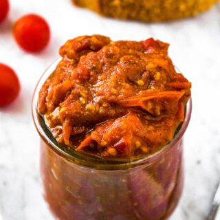 Close up overhead shot of a clear glass jar of grape tomato jam with a slice of rustic bread and tomato jam behind it