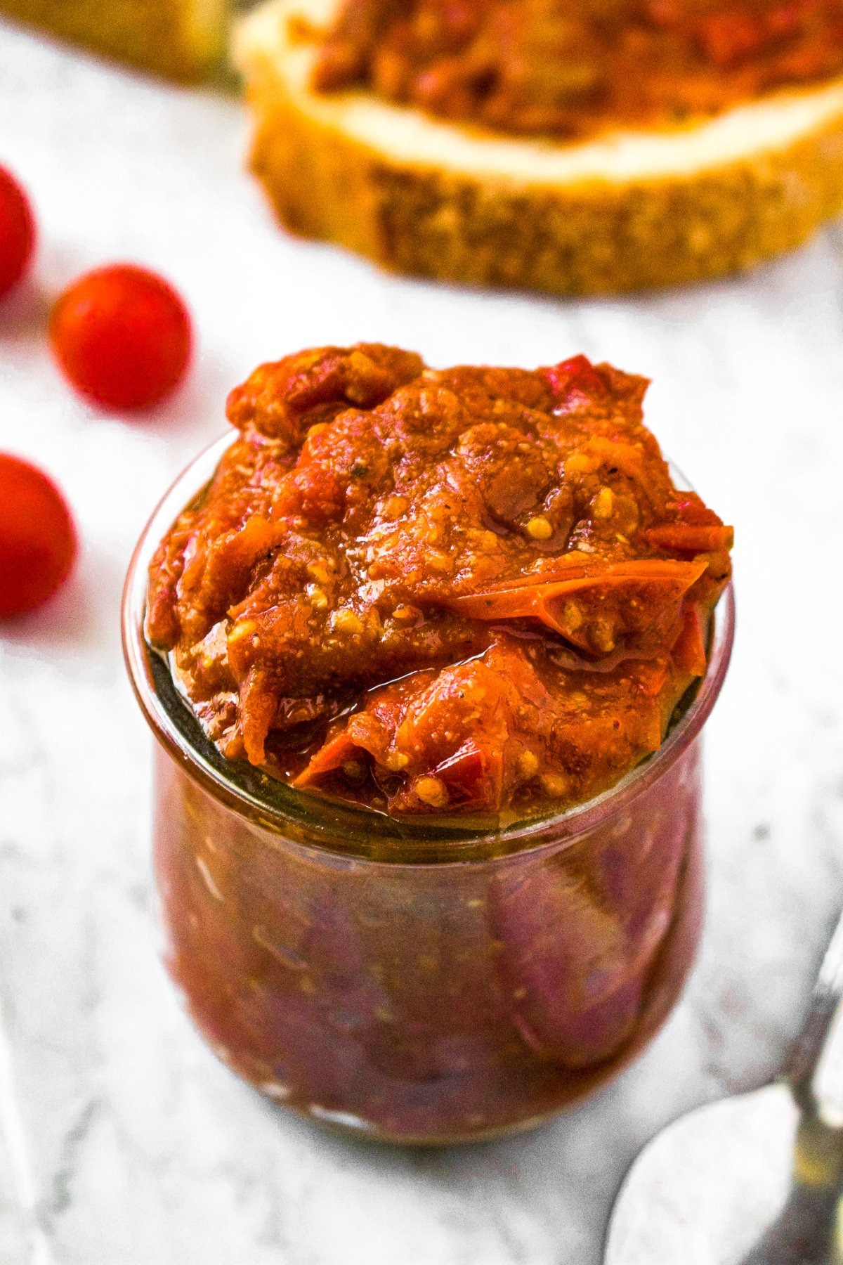 Close up overhead shot of a clear glass jar of grape tomato jam with a slice of rustic bread and tomato jam behind it