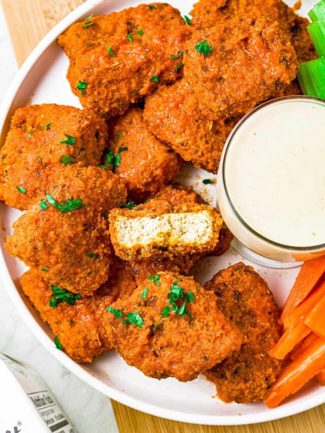Overhead photo of a pile of breaded and sauced vegan buffalo tofu wings with a bite taken out of the center wing to show the meaty texture inside.
