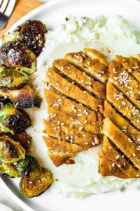 Overhead shot of a round white plate with mashed potatoes covering the bottom, roasted brussel sprouts to the left and on top, two honey garlic glazed tofu chicken cutlets sliced on a bias and sprinkled with sesame seeds