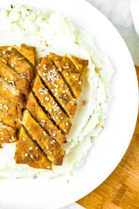 Overhead shot of a plate with a swirl of whipped potatoes topped with 2 honey soy tofu cutlets sliced on a bias and sprinkled with sesame seeds