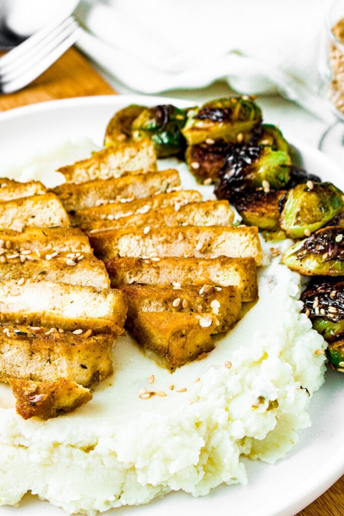 Head-on shot of a plate with mashed potatoes and brussels sprouts. On top of the mashed potatoes sits two sliced honey garlic tofu cutlets sprinkled with sesame seeds.