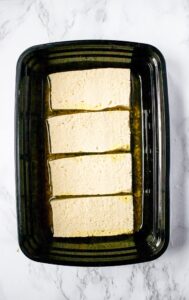 Overhead shot of a black rectangular container filled with four tofu cutlets on top of a layer of marinade