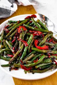 Overhead shot of a pile of green beans and red peppers on a round white plate with a large silver serving spoon to the top right