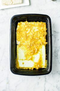 Overhead shot of two large squares of tofu in a black rectangular take-out container smothered in vegan schnitzel marinade