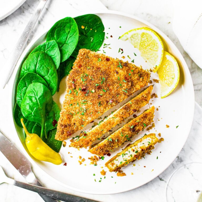 Overhead view of a vegan chicken cutlet, partially sliced, on a plate with fresh spinach leaves, lemon wedges, and pepporincini.