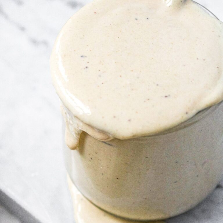 Close up overhead view of vegan tahini sauce for falafel overflowing in a glass jar with a spoon in it.