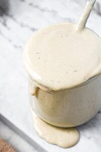 Close up overhead shot of vegan tahini sauce overflowing in a glass jar with a spoon in it.