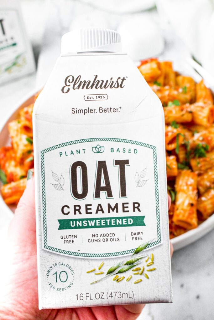 Overhead shot of a hand holding a carton of Elmhurst's unsweetened oat creamer over a plate of rigatoni