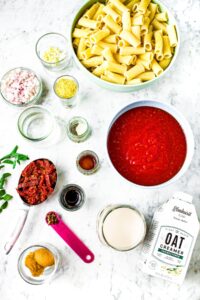 Overhead shot of all the ingredients you need to make vegan vodka pasta: crushed tomatoes, pasta, sun-dried tomatoes, shallots, garlic, nutritional yeast miso paste, liquid smoke, soy sauce, Elmhurst unsweetened oat creamer, vodka, and spices