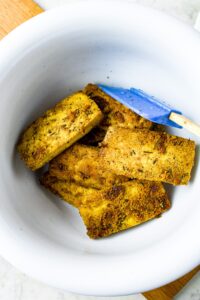 Overhead shot of baked tofu slices in a white mixing bowl with a blue rubber spatula stirring them
