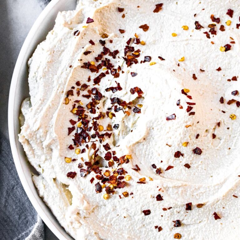 Close up overhead view of a circular schmear of dairy-free ricotta on a round white plate sprinkled with red pepper flakes and surrounded by a gray linen