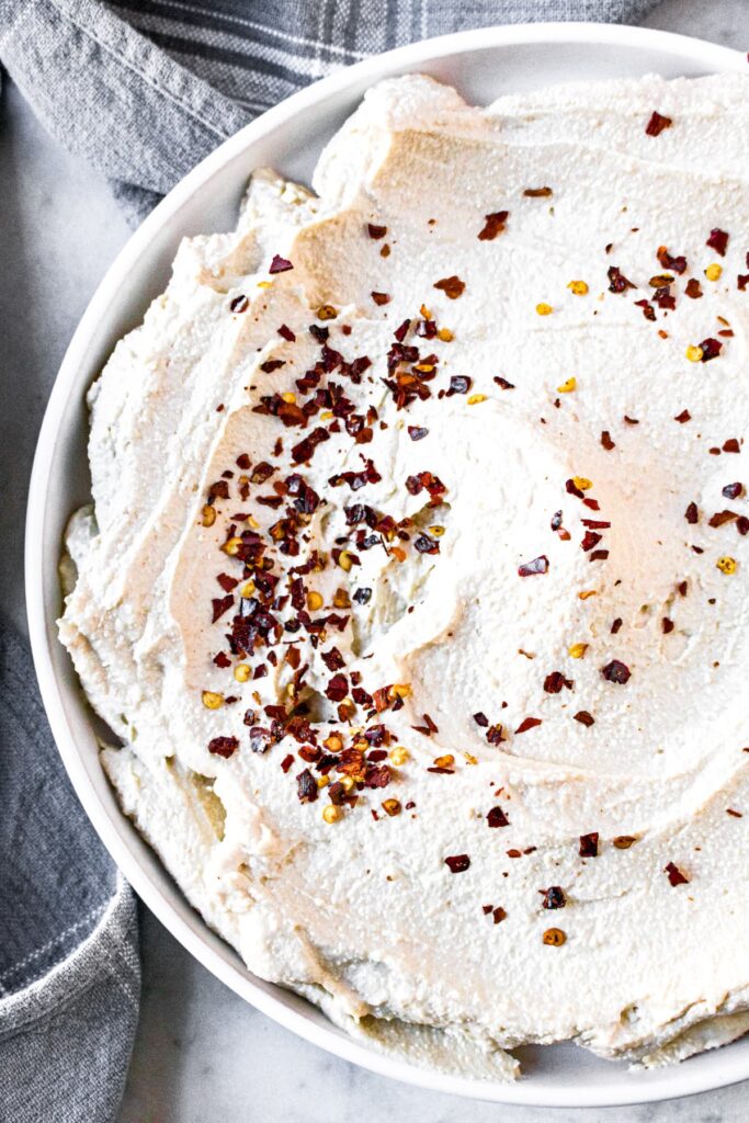 Close up overhead shot of a circular schmear of dairy-free ricotta on a round white plate sprinkled with red pepper flakes and surrounded by a gray linen