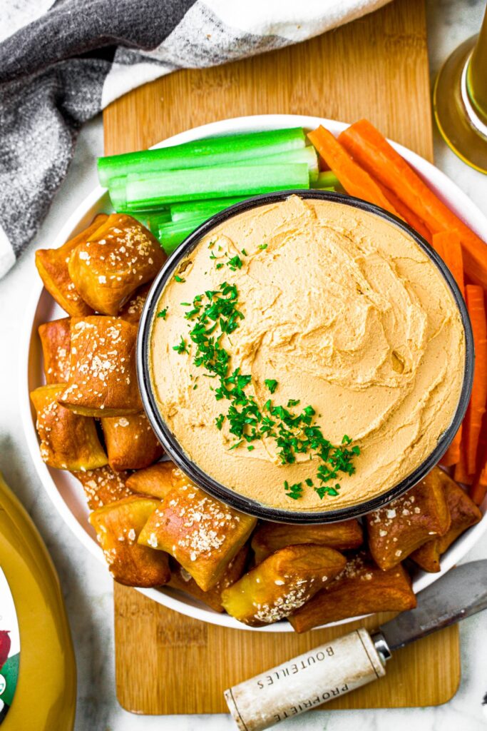 Overhead shot of a bowl of plant-based pub cheese spread garnished with fresh minced herbs and surrounded by celery and carrot sticks and soft pretzel bites