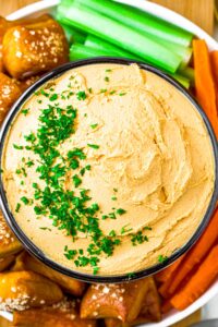 Overhead zoomed in shot of a bowl of vegan beer cheese dip topped with fresh minced herbs and surrounded by celery and carrot sticks and soft pretzel bites