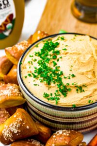 Close up head on shot of a bowl of thick tofu beer cheese topped with minced green parsley and surrounded by mini soft pretzel bites