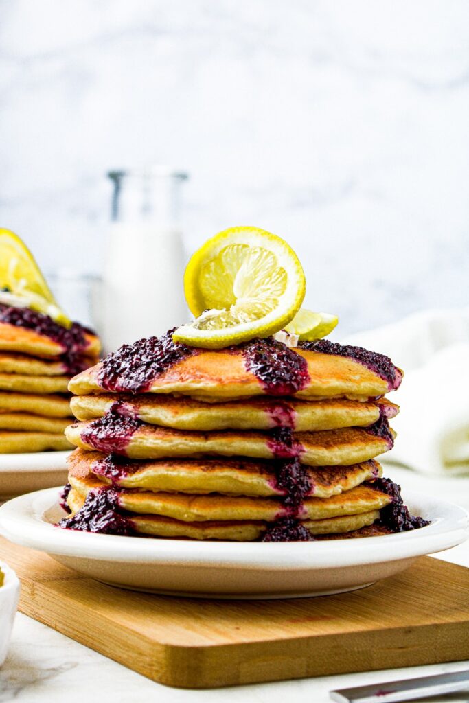Head on shot of a stack of vegan ricotta pancakes with blueberry compote dripping over the edges and a twisted lemon slice on top