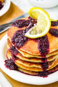 Overhead shot of a stack of lemon ricotta pancakes with blueberry syrup dripping down the edges and a twisted lemon slice sitting on top