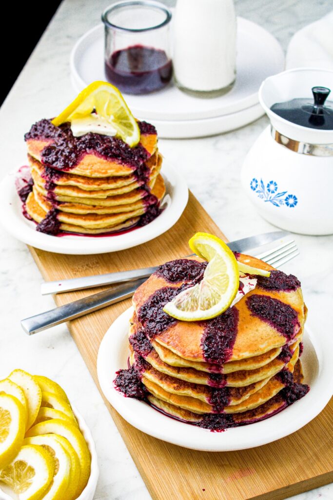 Overhead shot of two small plates with stacks of lemon ricotta pancakes topped with blueberry sauce and twirly lemon slices. There is a small coffee pot in the top right corner and lemon slices in the bottom right corner