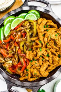 Close up photo of a round black cast iron pan filled with seared soy curls, red and green peppers and onions, and lime wedges