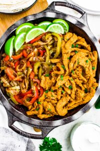 Overhead close up photo of a deep cast iron pan filled with vegan chicken, peppers and onions, and limes for fajitas