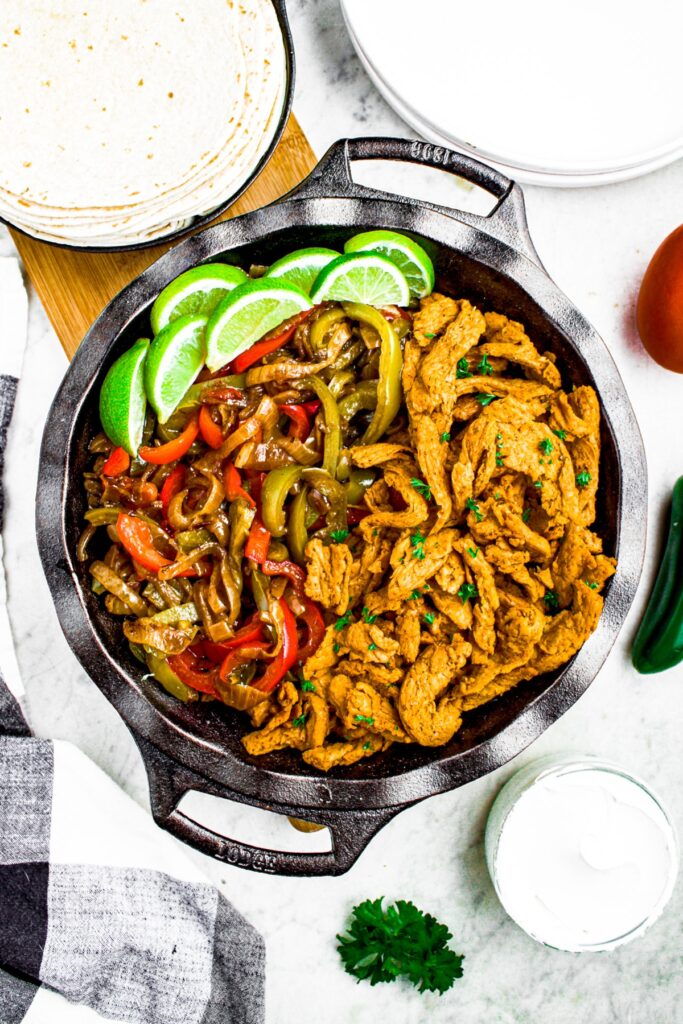 Overhead shot of a cast iron pan filled with vegan fajita fillings: soy curls, peppers and onions, and lime wedges