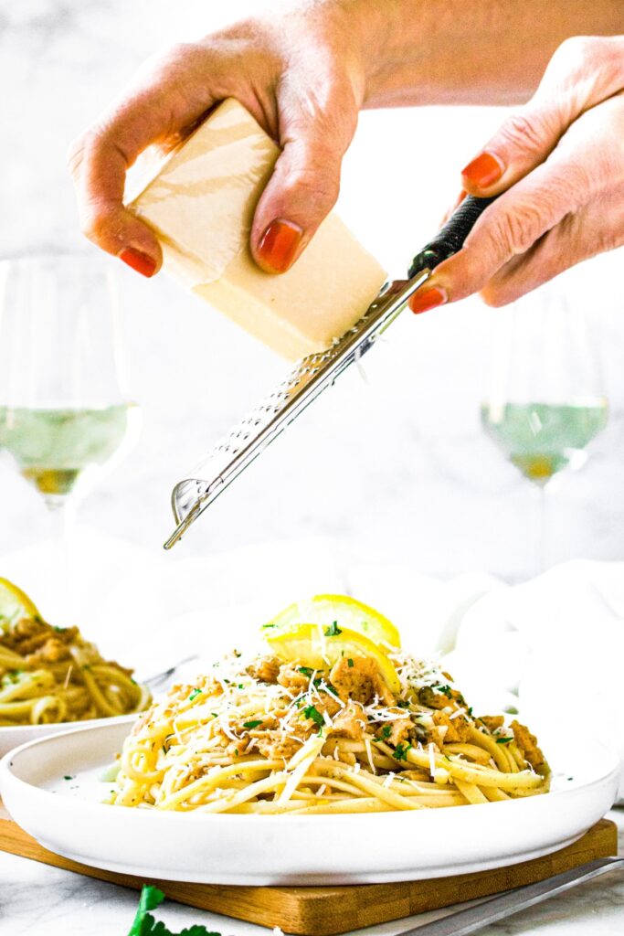 Head on shot of a pile of vegan clam sauce pasta on a plate with hands overhead grating dairy-free parmesan on the dish. Two glasses of white wine can be seen in the background.