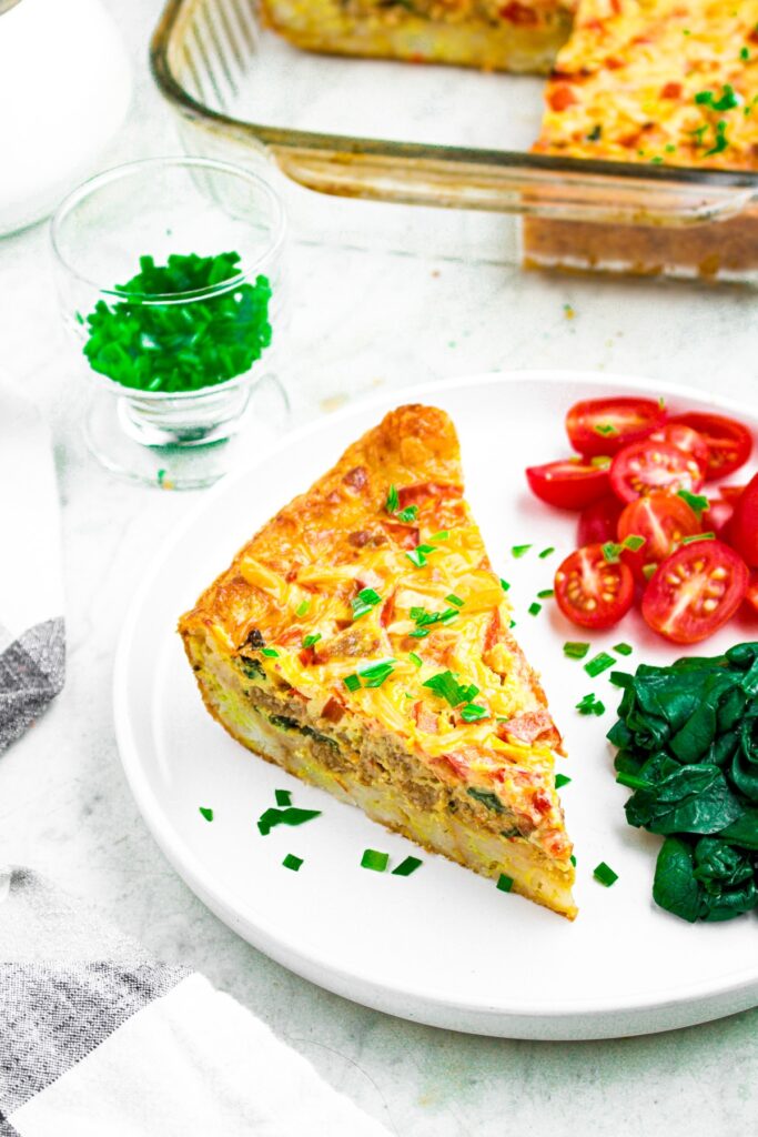 Overhead photo of a triangular slice of vegan breakfast casserole sprinkled with fresh herbs on a plate with tomatoes and spinach